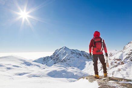 picture of a person in a red hooded jacket and black pants on top of a snowy mountain with a bright starburst sun and a clear blue sky