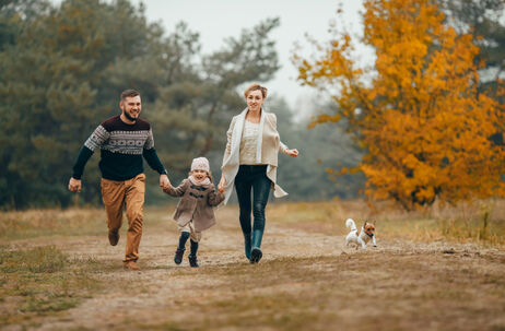 photo of a dad, mom, and toddler holding hands and running down a path in the fall while their dog runs beside them