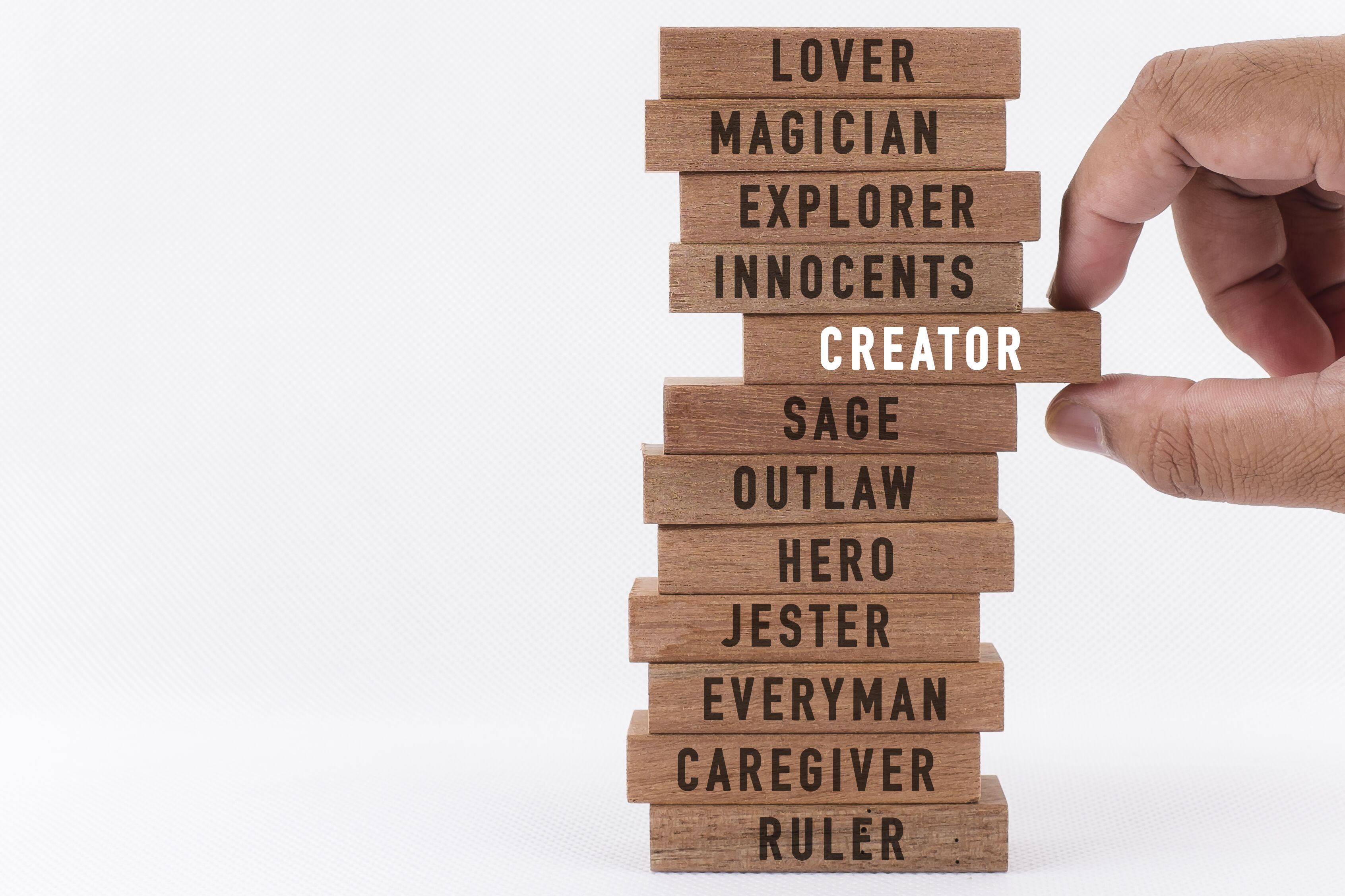 wooden blocks stacked on top of each other with the words lover, magician, explorer, innocents, creator, sage, outlaw, hero, jester, everyman, caregiver, and ruler written individually on each. The creator block is being pulled out from the middle by a person's thumb and forefinger