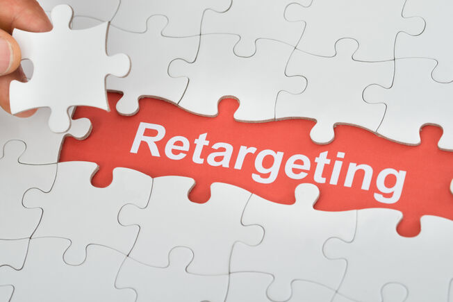 An all white puzzle that is missing a few pieces to reveal the word "retargeting" written in white on a red background, with some fingertips holding a single white puzzle piece