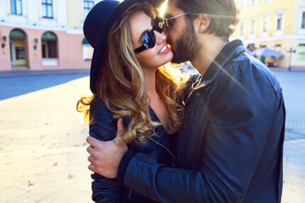 photo of a couple who are both wearing black clothes and sunglasses on a street where the woman is wearing a black hat and the man is grabbing her by the shoulders and speaking in her ear