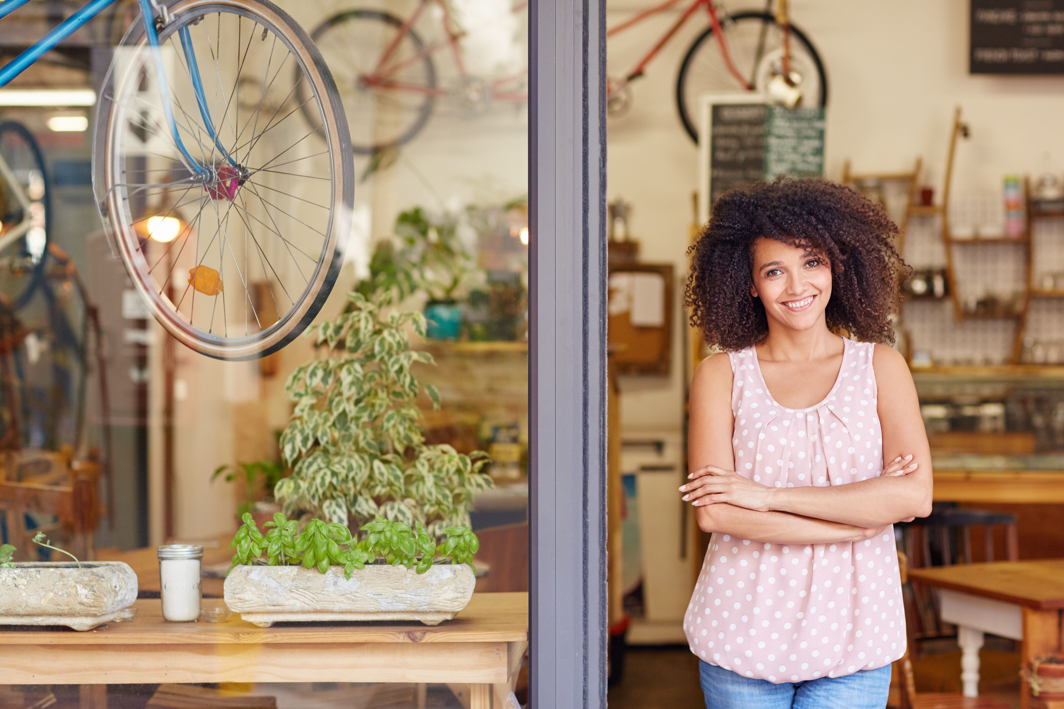 young woman standing in the doorway of a cute shop with plants in the window smiling with arms folded in front of her