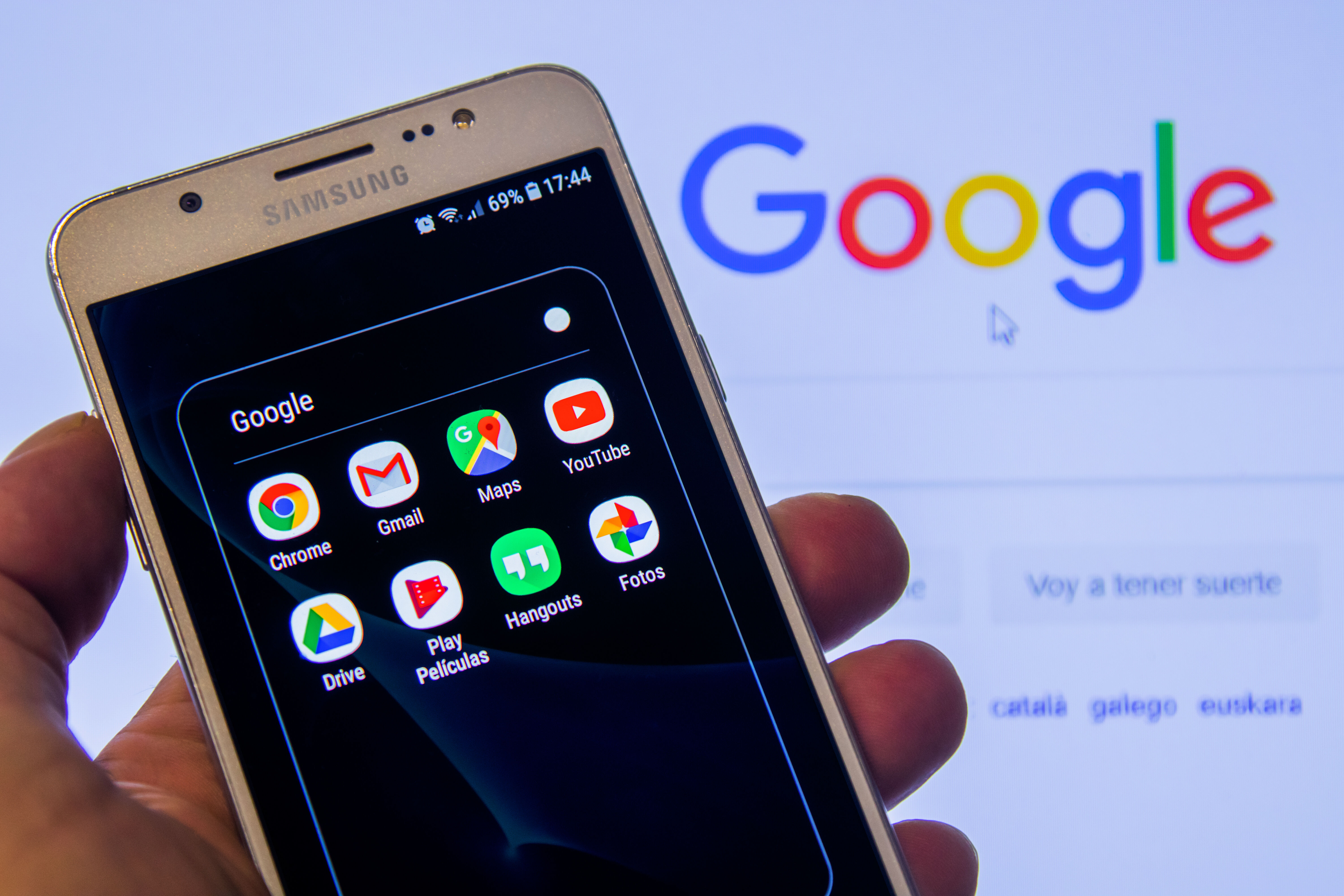google knows everything about you hand holding a samsung smart phone with the google apps icons on the phone's screen, with a google search homepage in the background