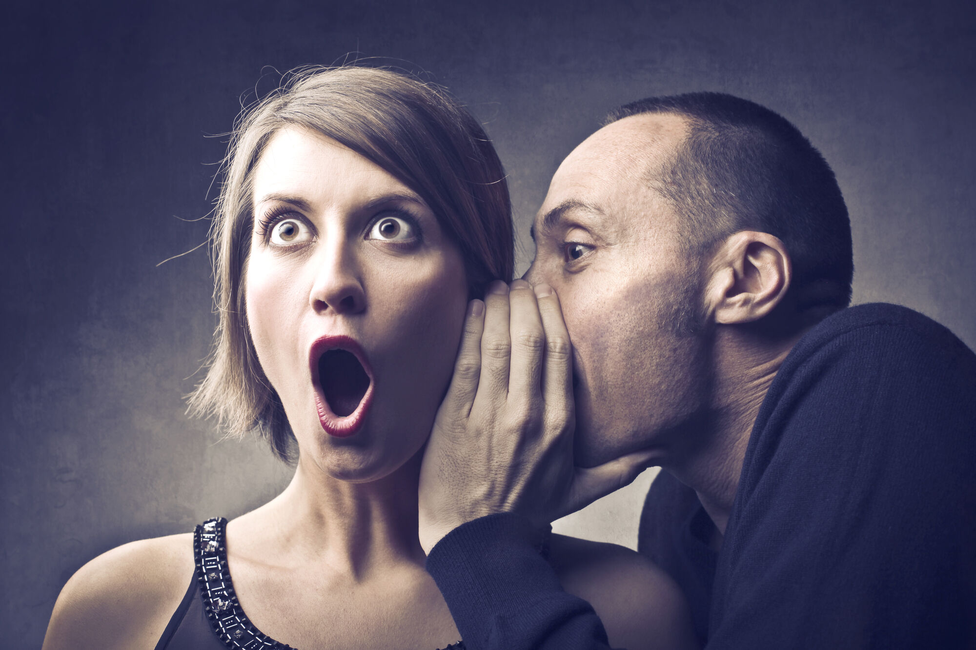 Man whispering a secret to surprised woman, her eyes wide and mouth open in shock