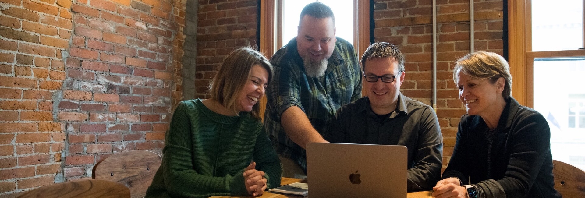 Four people smile and collaborate over a laptop in a cozy, brick-walled setting.