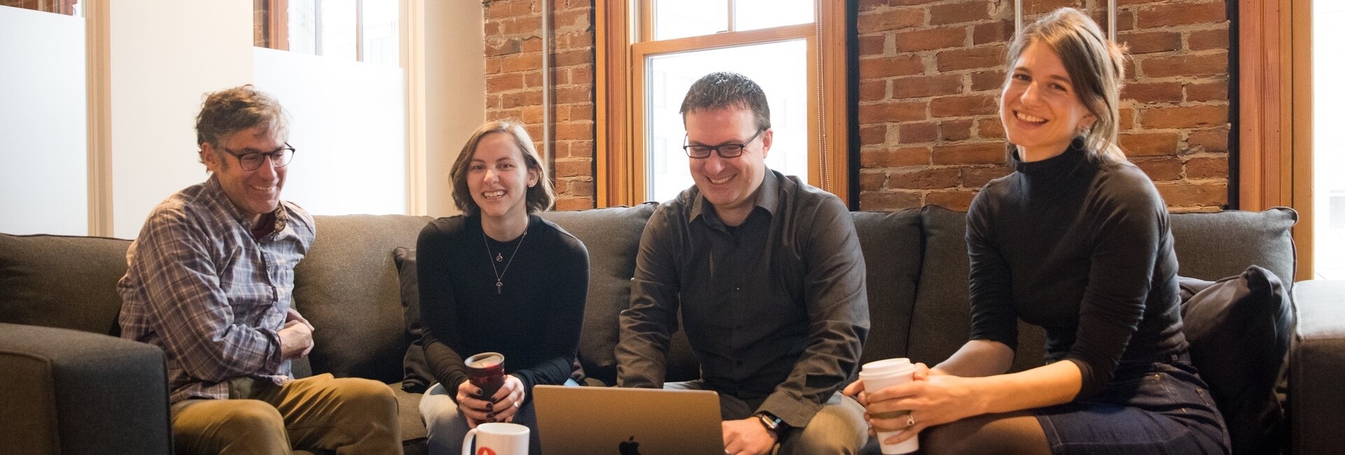 Four people are smiling and engaged around a laptop on a couch in a cozy setting.