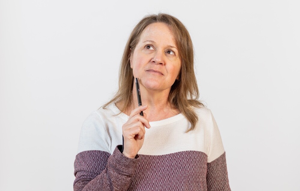 Holly Buttura looking upward in thought with a pen in hand against a plain white background.