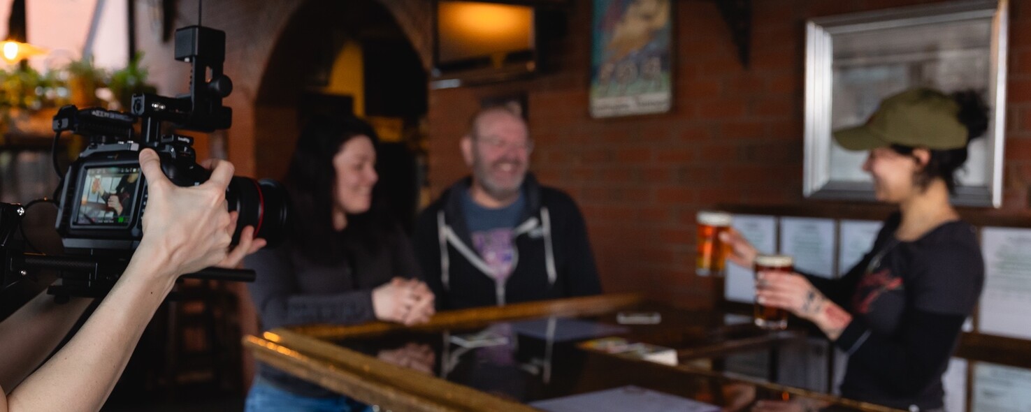 A bartender serves drinks to two smiling customers in a cozy pub setting.