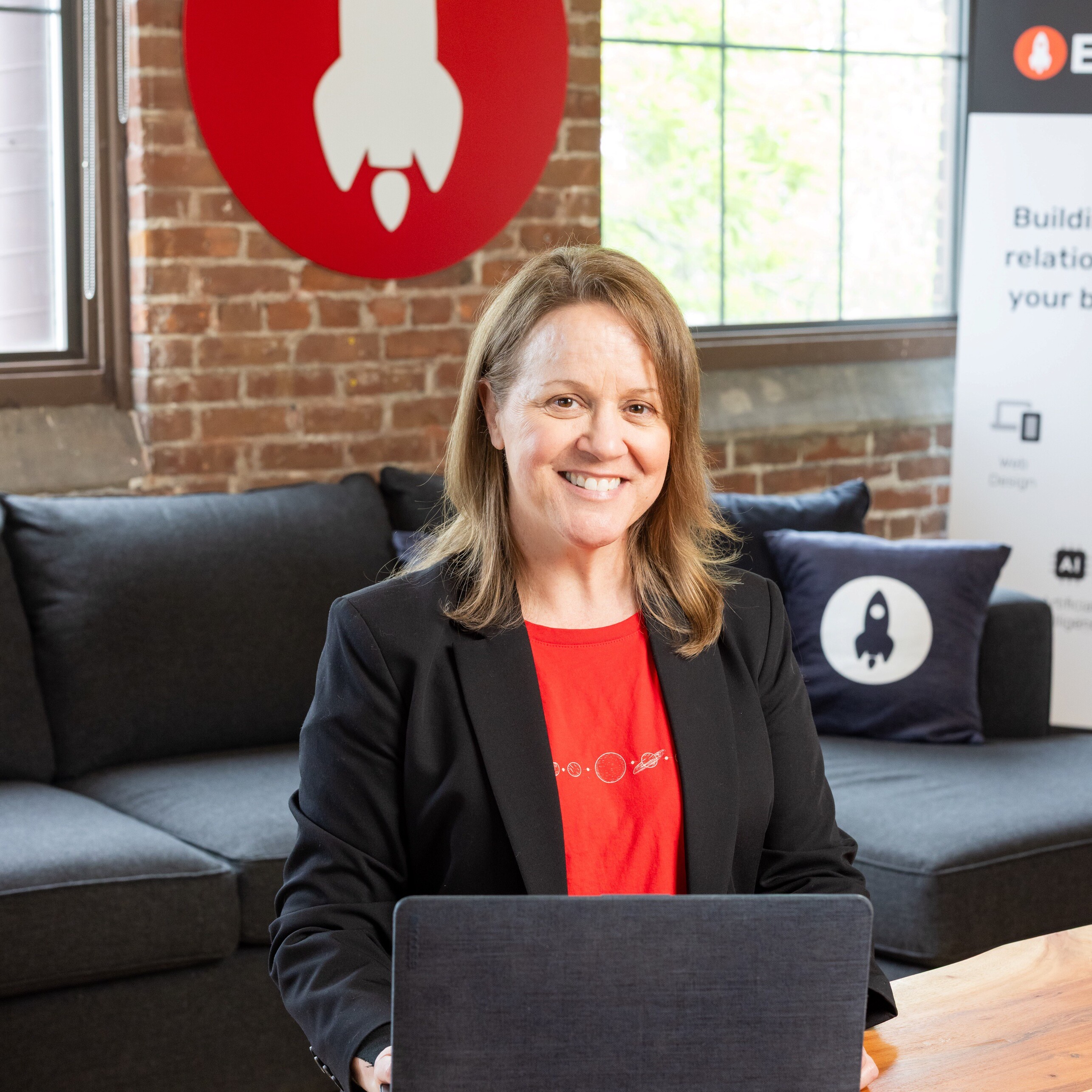 Smiling woman in a blazer at a laptop, with a rocket logo backdrop.