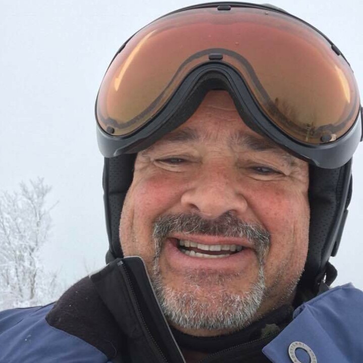 Smiling man wearing ski gear and goggles in a snowy landscape.