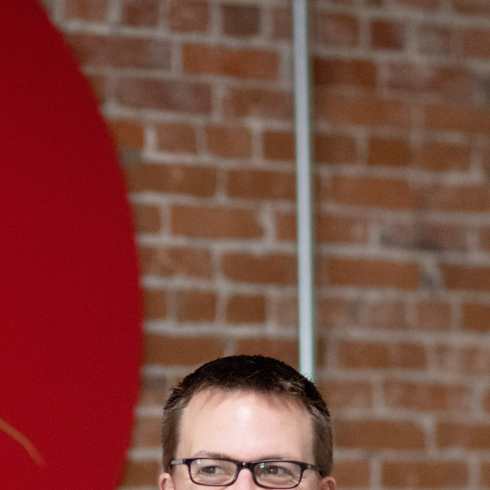 Person with glasses in front of a red circle, against a brick wall background.