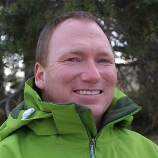 Smiling man in a green jacket outdoors, with trees in the background.