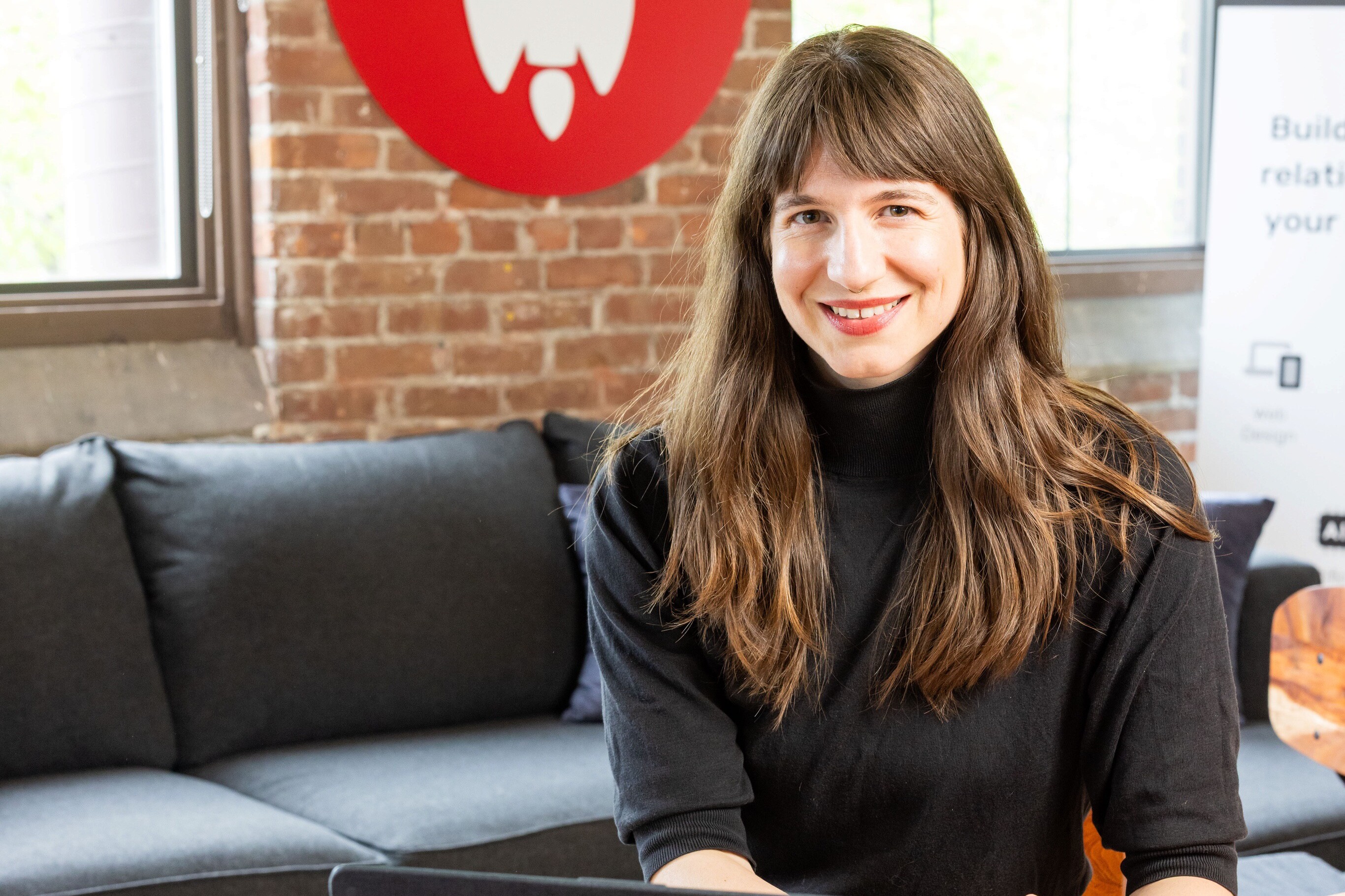 A smiling woman in a black sweater sitting at a laptop in a cozy, modern workspace.