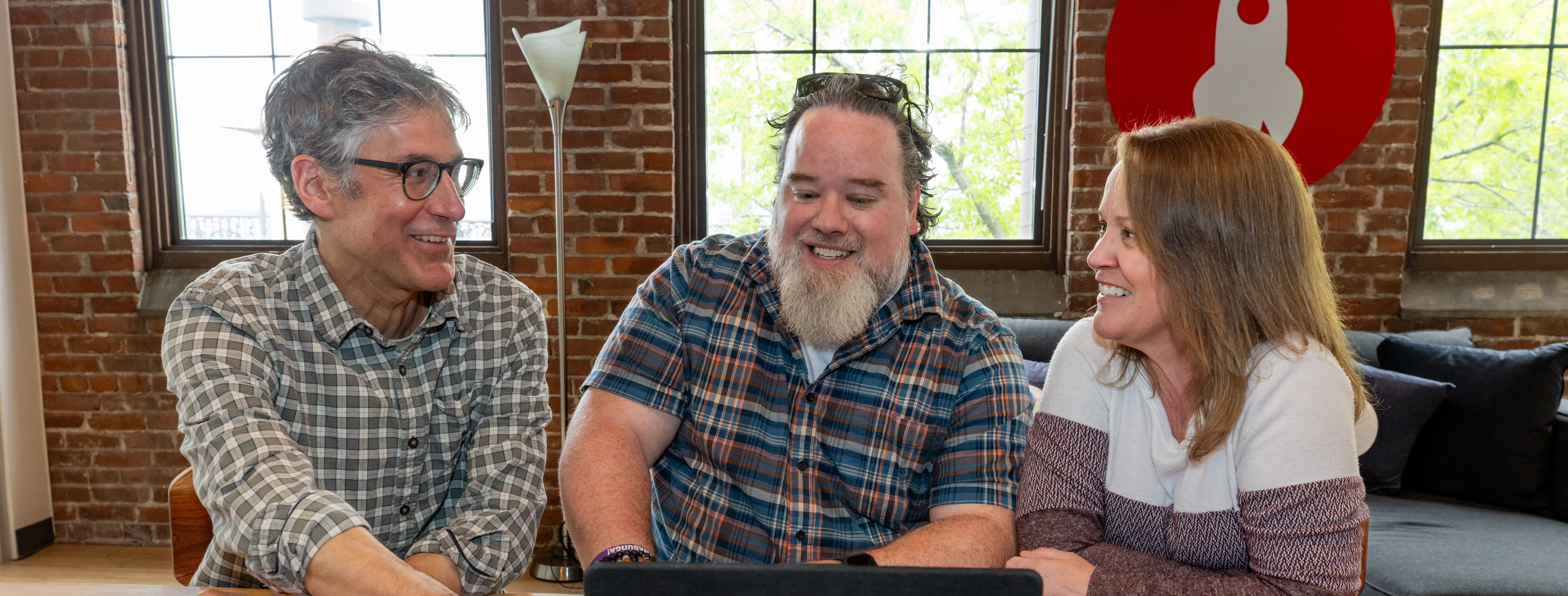 Three people discuss something on a laptop in a cozy, brick-walled setting.