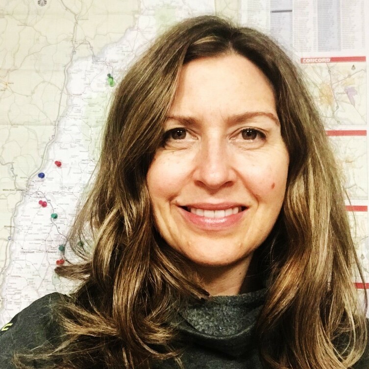 Woman with long hair smiles in front of a map covered with colorful pins.