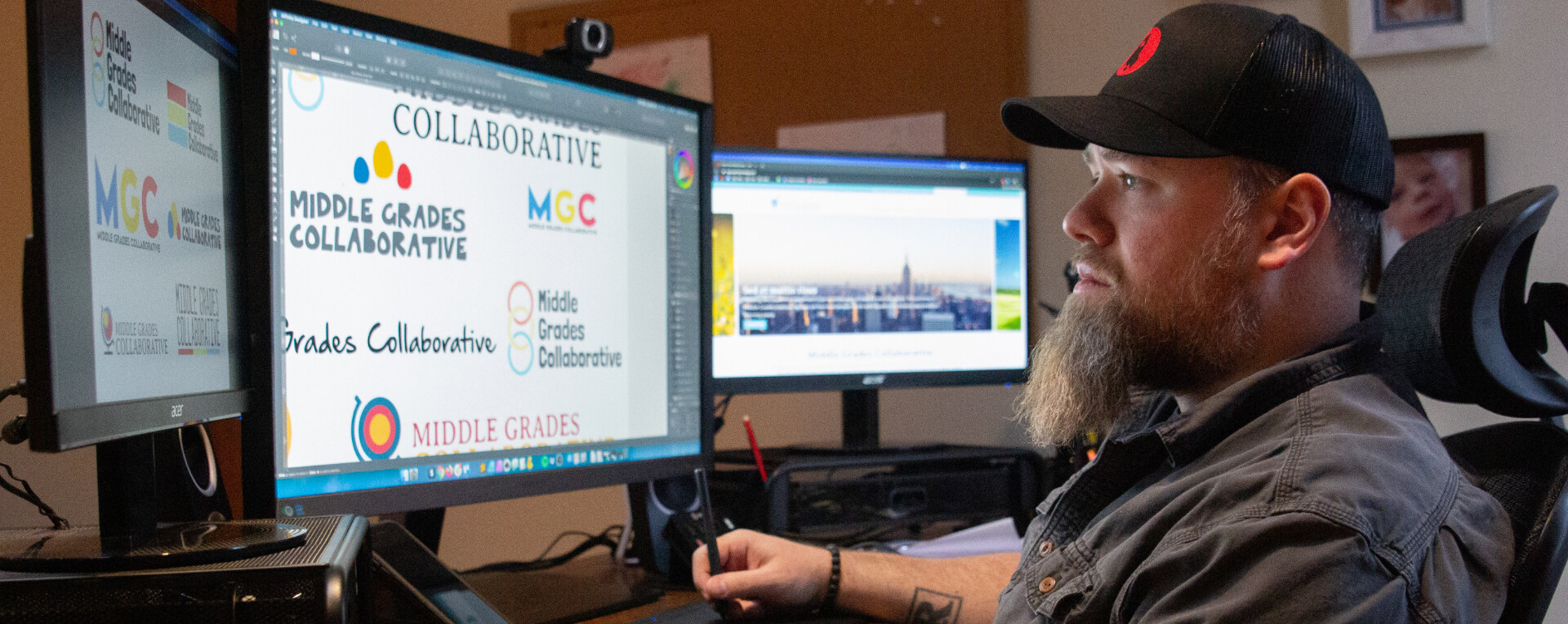 Person working at a desk with dual monitors displaying logos for Middle Grades Collaborative.