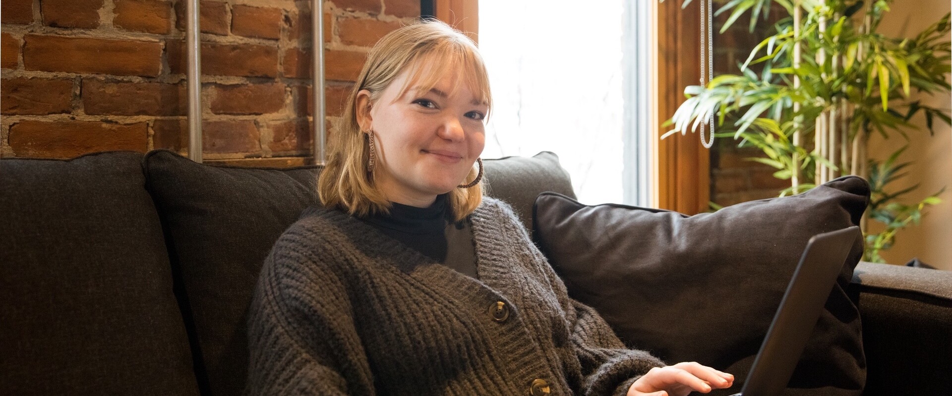 Young woman smiling on a couch, using a laptop in a cozy, brick-walled setting.