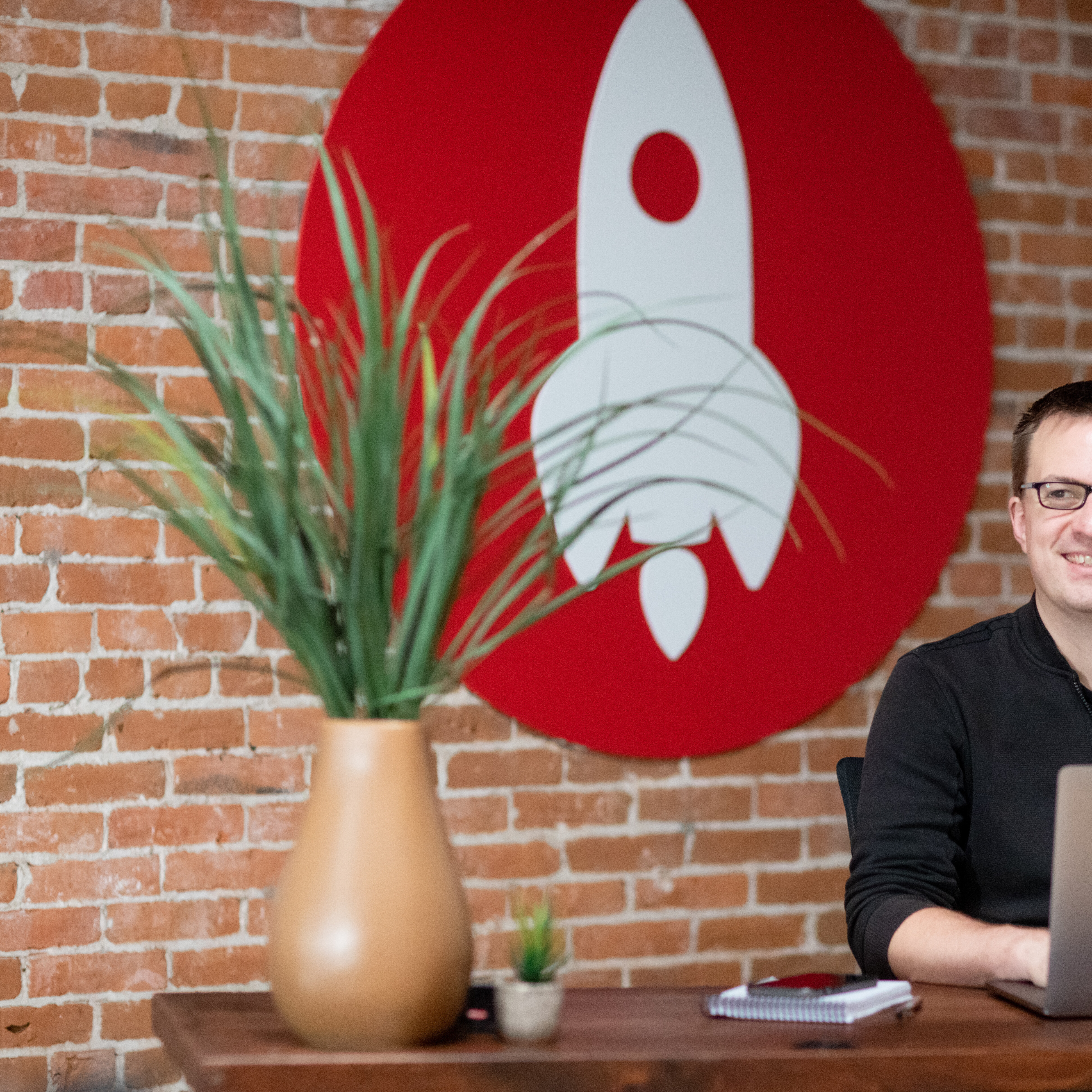Person sitting at a desk in front of a rocket-themed wall with a plant nearby.