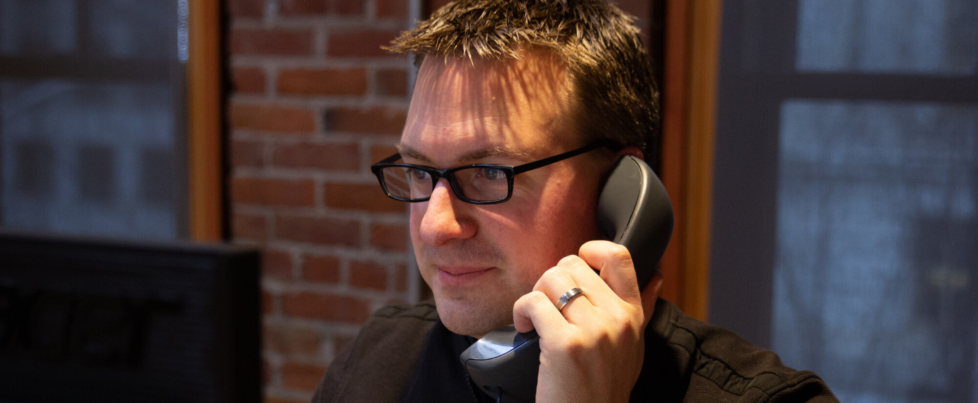 Person with glasses on the phone in a brick-walled office setting.