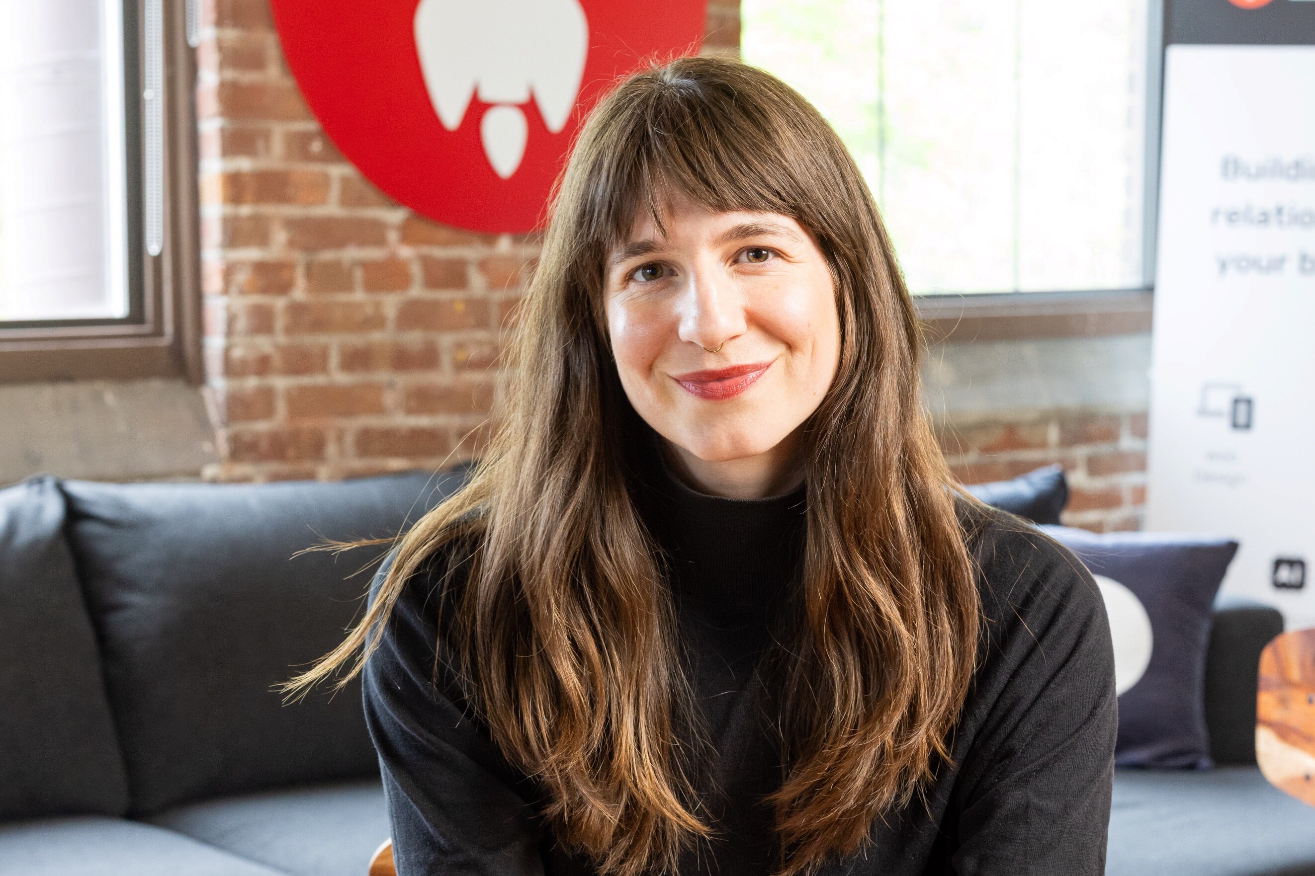 A woman with long hair smiles warmly, sitting on a couch in a modern workspace.