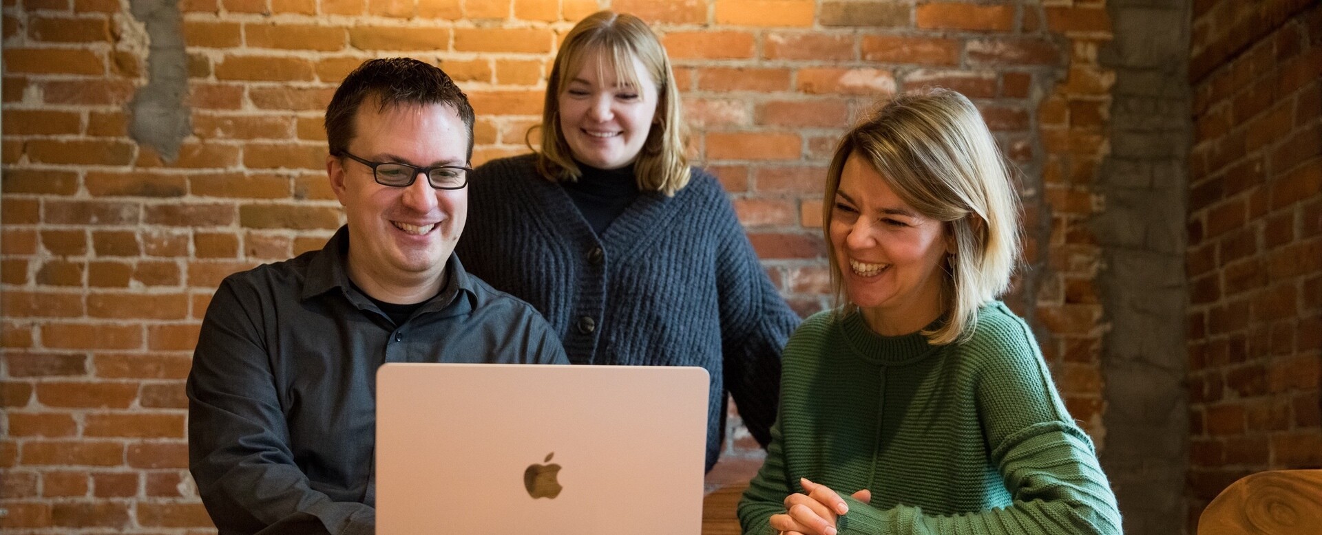 Three people smiling at a laptop in a cozy, brick-walled workspace.