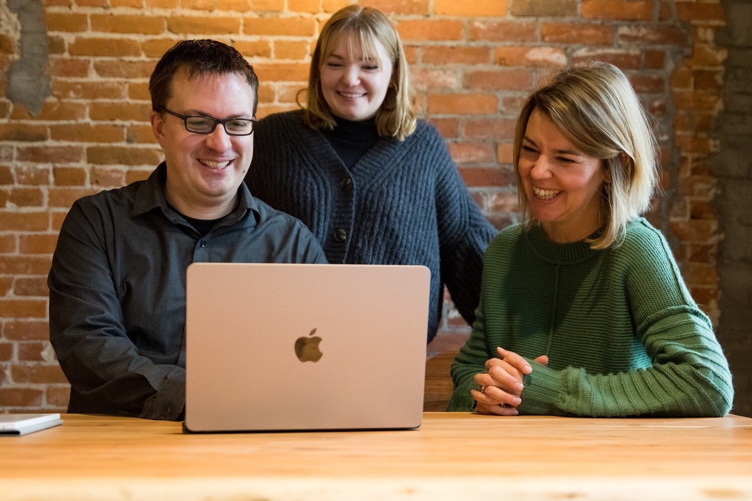 Three people are smiling and looking at a laptop on a wooden table in a cozy setting.