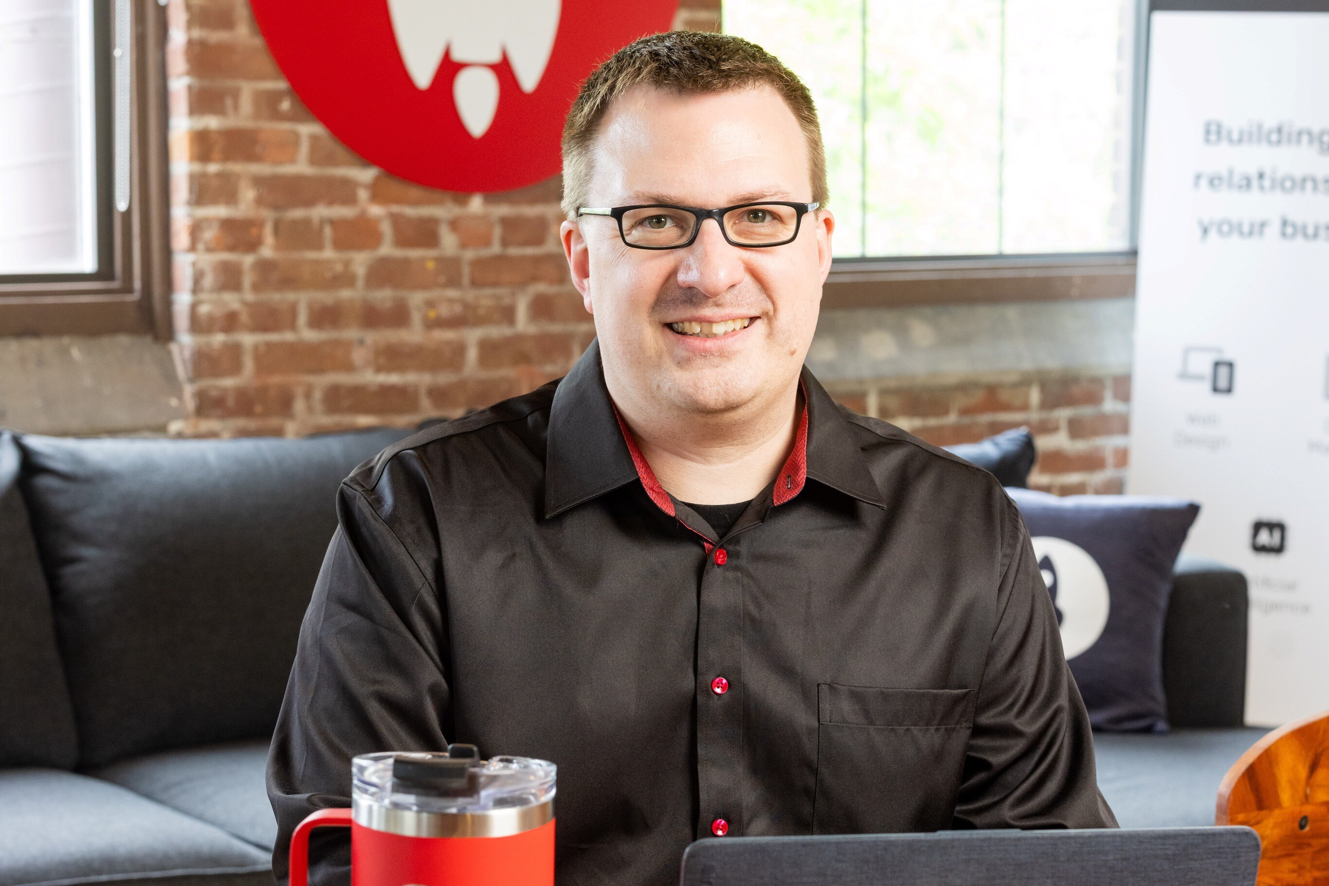 A smiling man in a black shirt is sitting at a desk with a drink and laptop.