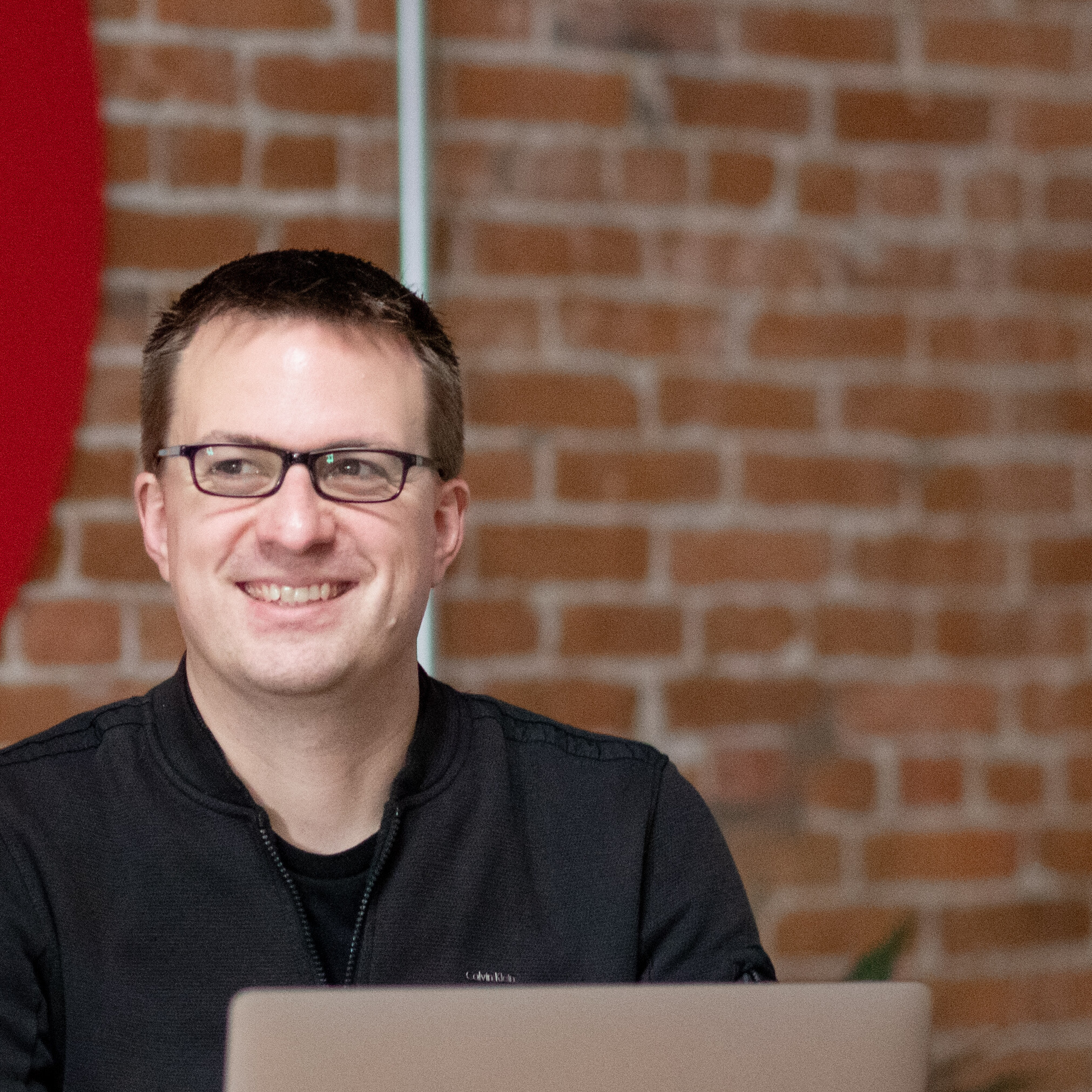 Smiling person in glasses sits at a laptop against a brick wall background.