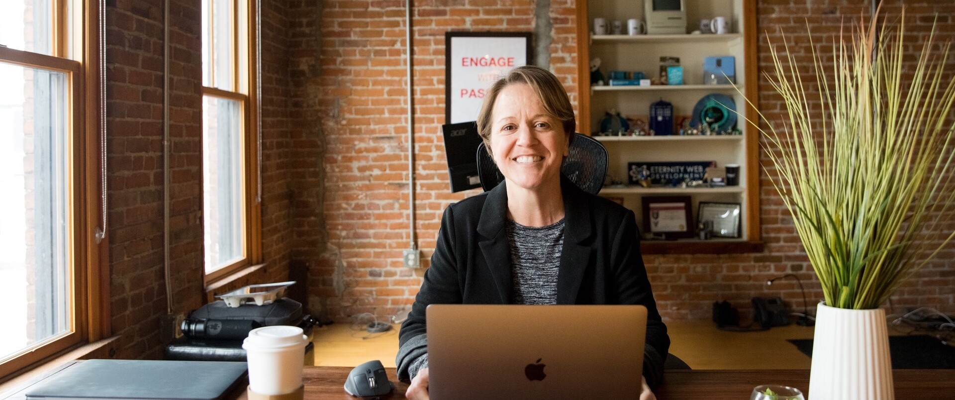 Person smiling at a desk in a brick office, with a laptop and plants nearby.