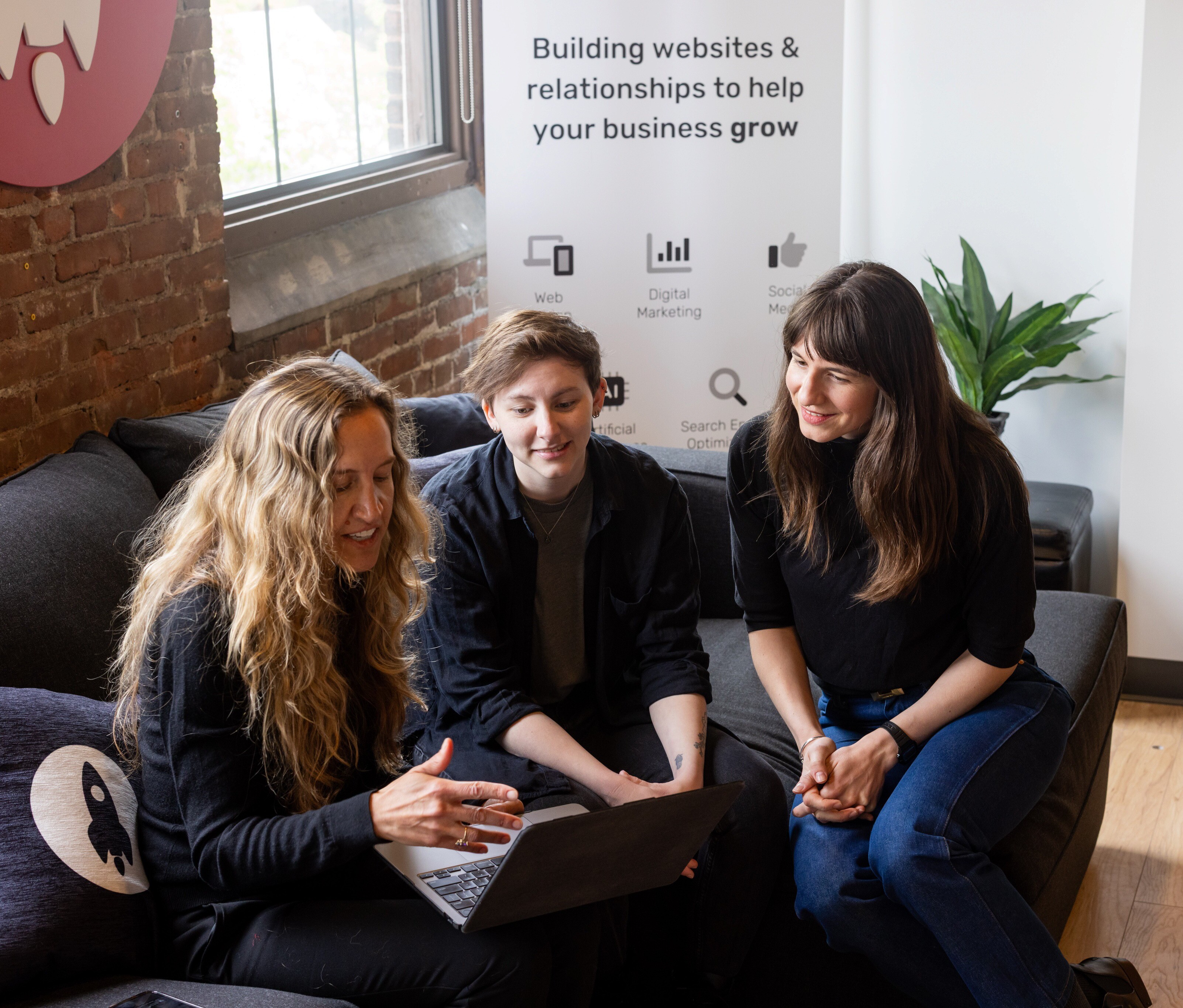 Three women collaborate on a laptop in a casual workspace with a banner in the background.