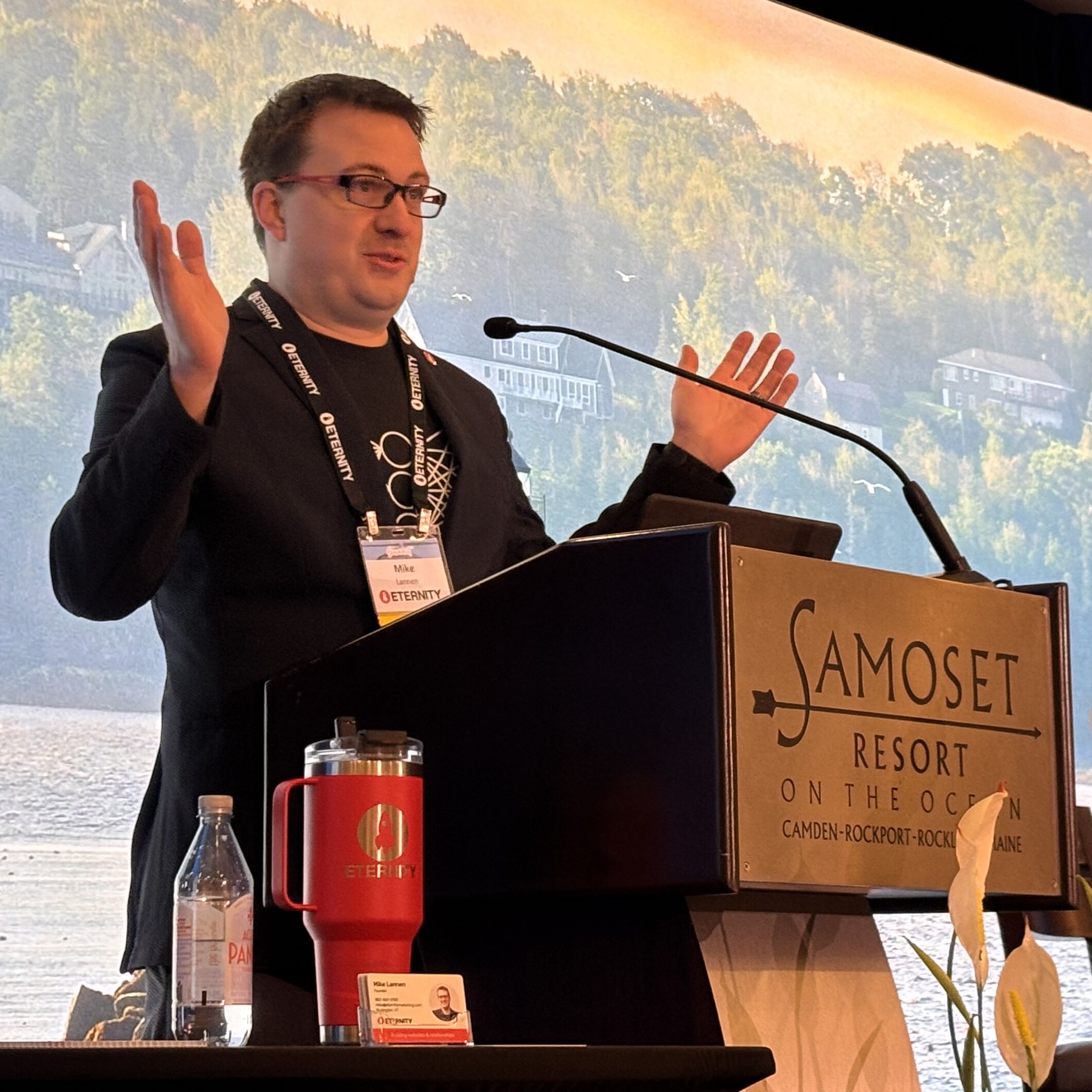 Speaker presenting at a podium with a scenic background and refreshments on the table