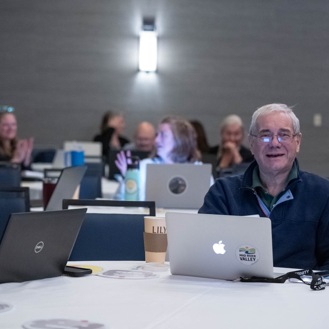 A smiling man sits at a table with laptops and coffee cups during a conference or event.