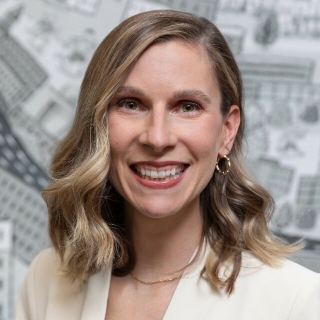 Person smiling in front of a patterned background, wearing a white blazer and earrings.