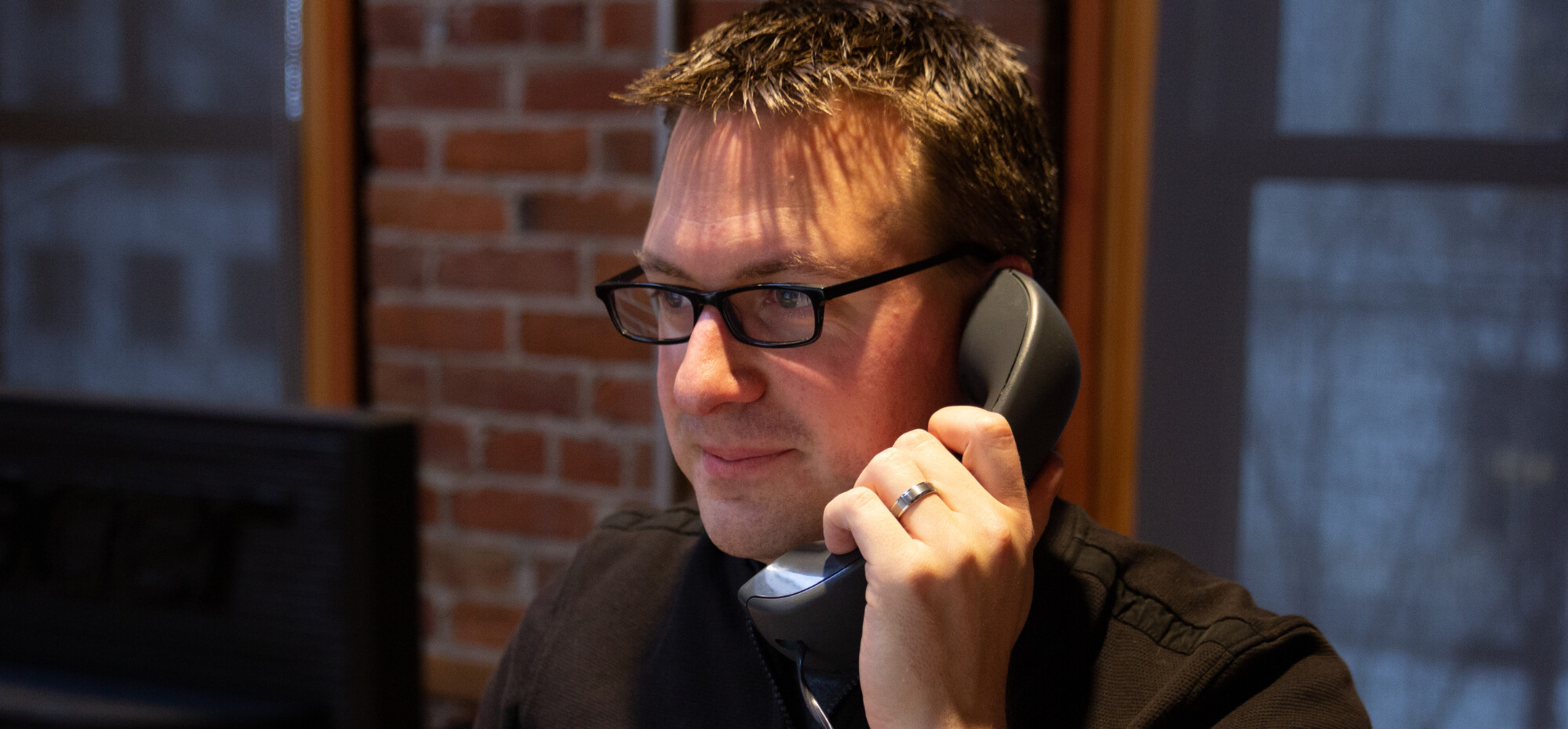Person with glasses talking on a phone in a brick-walled office.