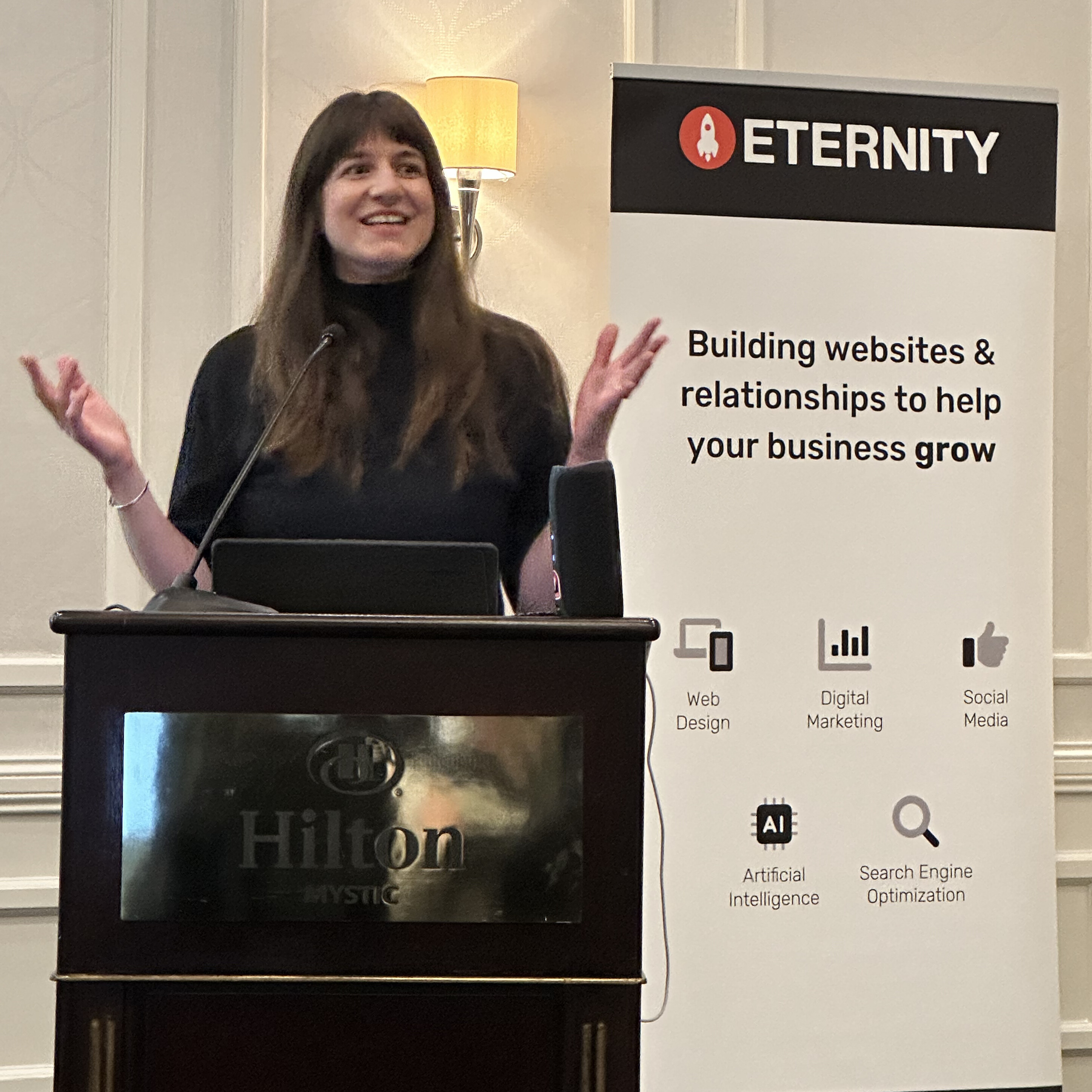 A woman speaks at a podium beside a banner for Eternity, discussing business growth.