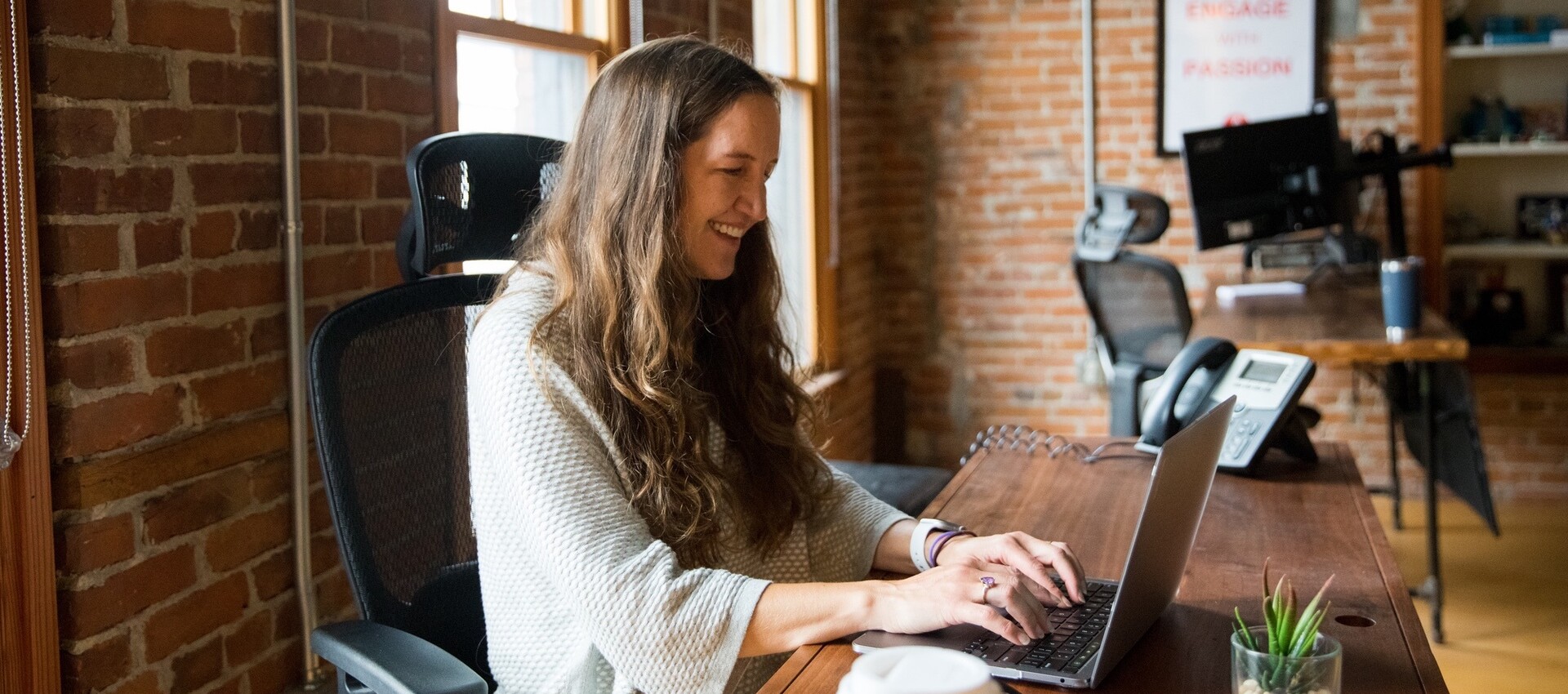 A woman smiles while typing on a laptop in a cozy office setting.