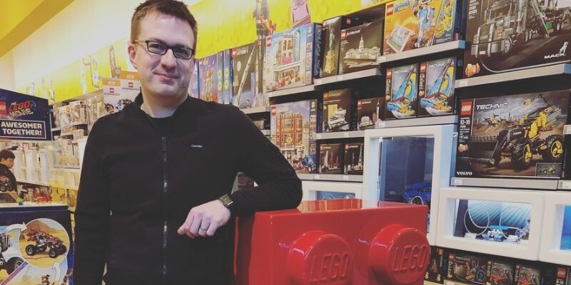 Mike Lannen stands next to a large LEGO brick in a toy store filled with LEGO sets.