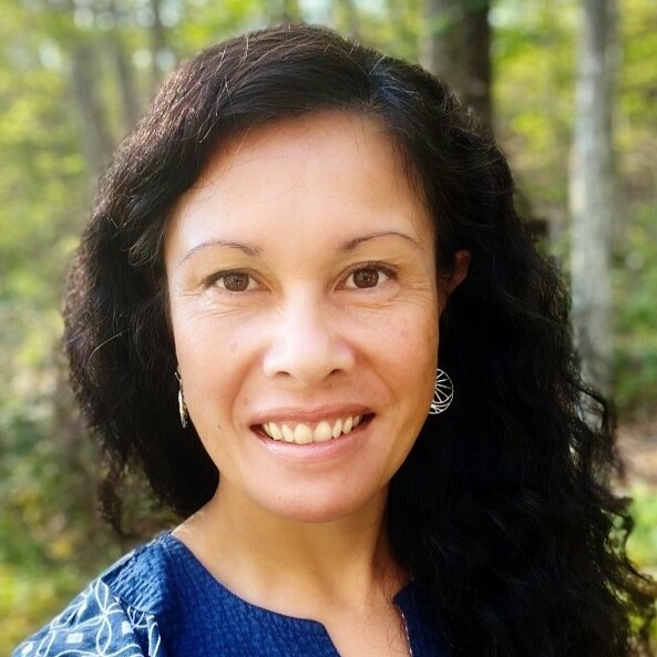 Smiling woman with curly hair outdoors, wearing a blue top and earrings.