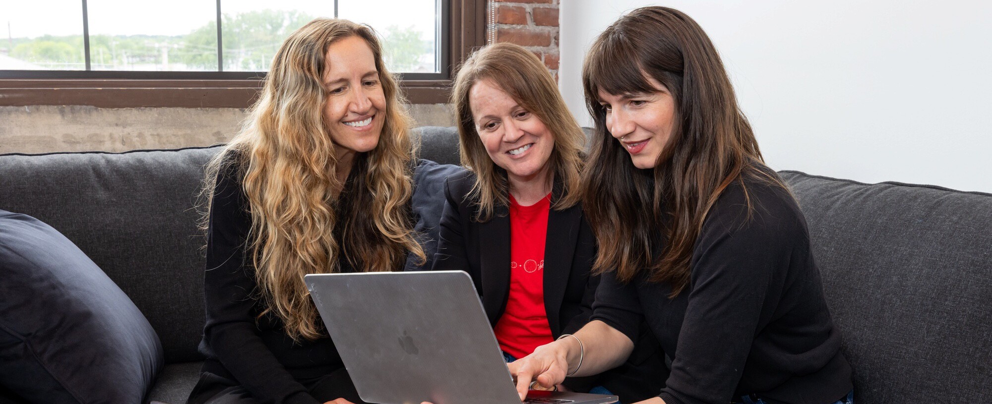 Three women collaborate, smiling and looking at a laptop while seated on a couch.