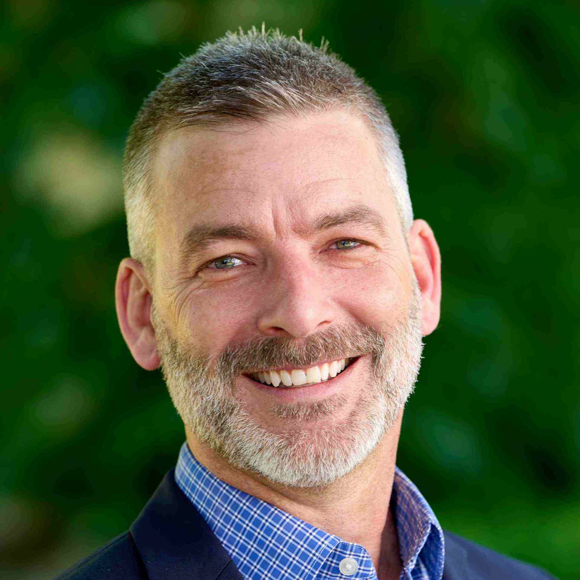 Smiling man with a short beard, wearing a blazer and checked shirt against a green background.
