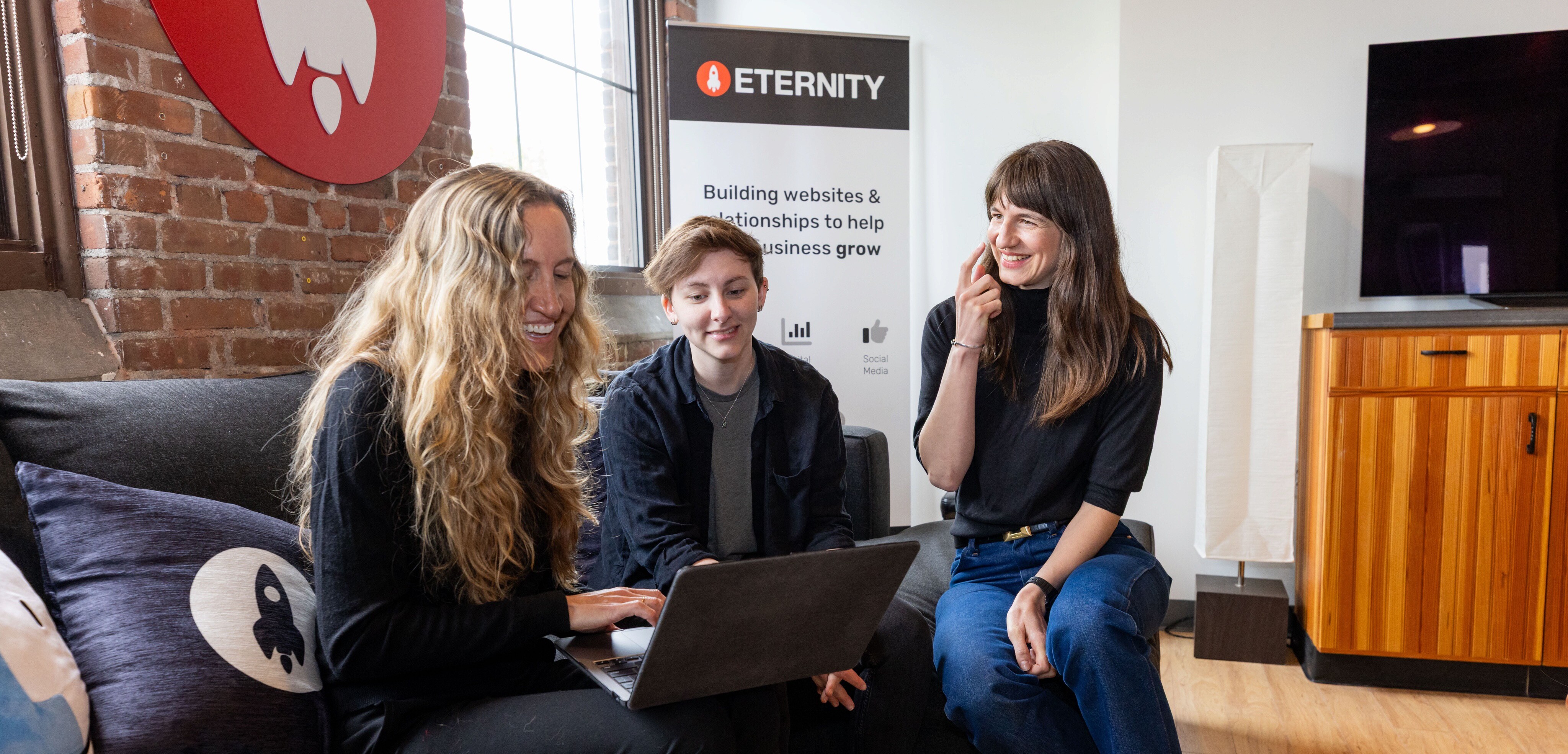 Three women collaborate on a laptop while seated on a couch, smiling and engaged.