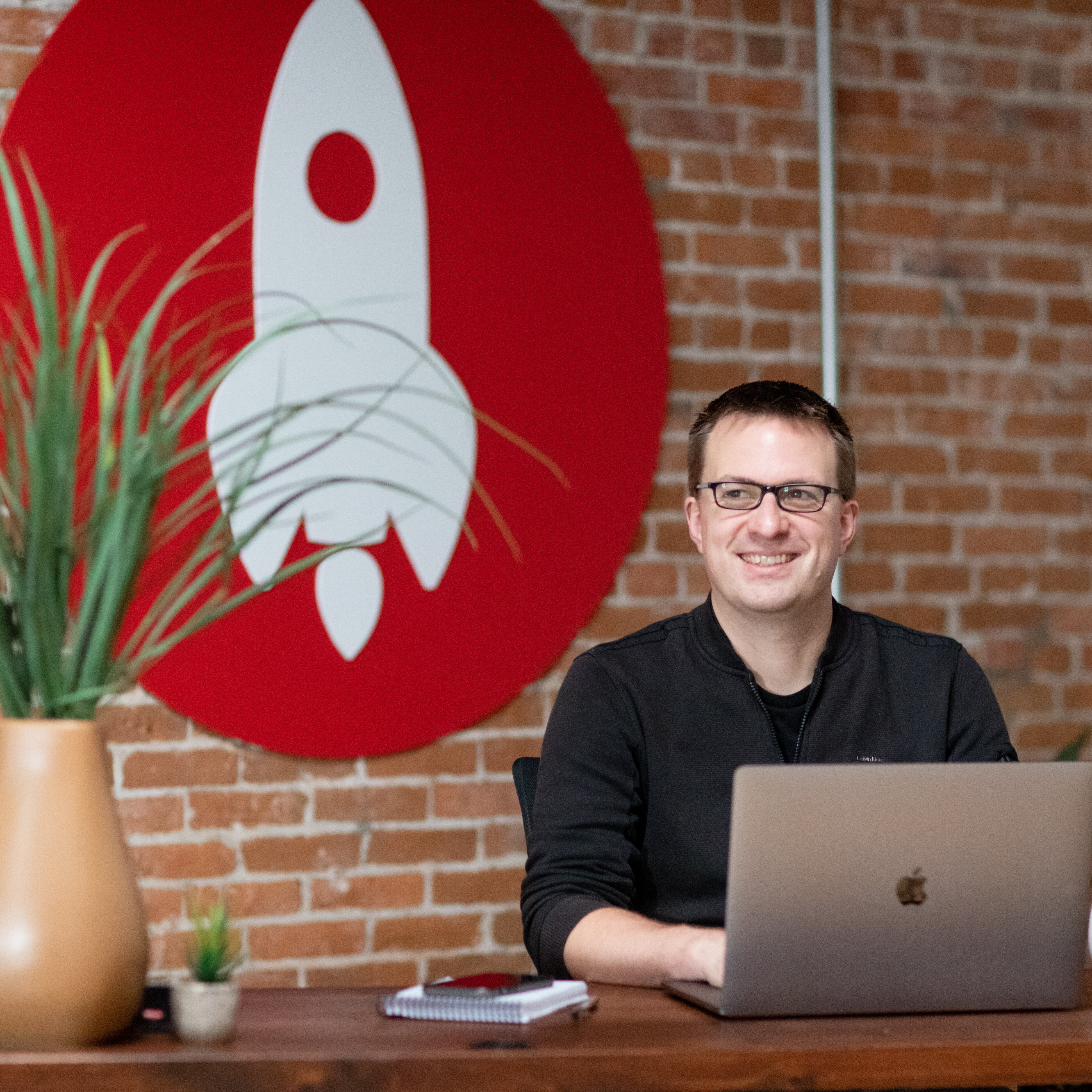 Smiling man at a desk with a laptop, plants, and a rocket-themed wall mural.