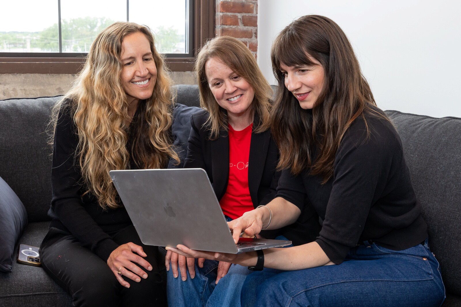 Jena Durell, Holly Buttura and Sophie Bodnar sit together on a couch, looking at a laptop and smiling.