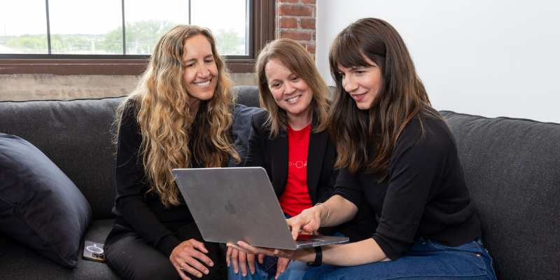 Three women smile while looking at a laptop on a couch in a bright, casual setting.