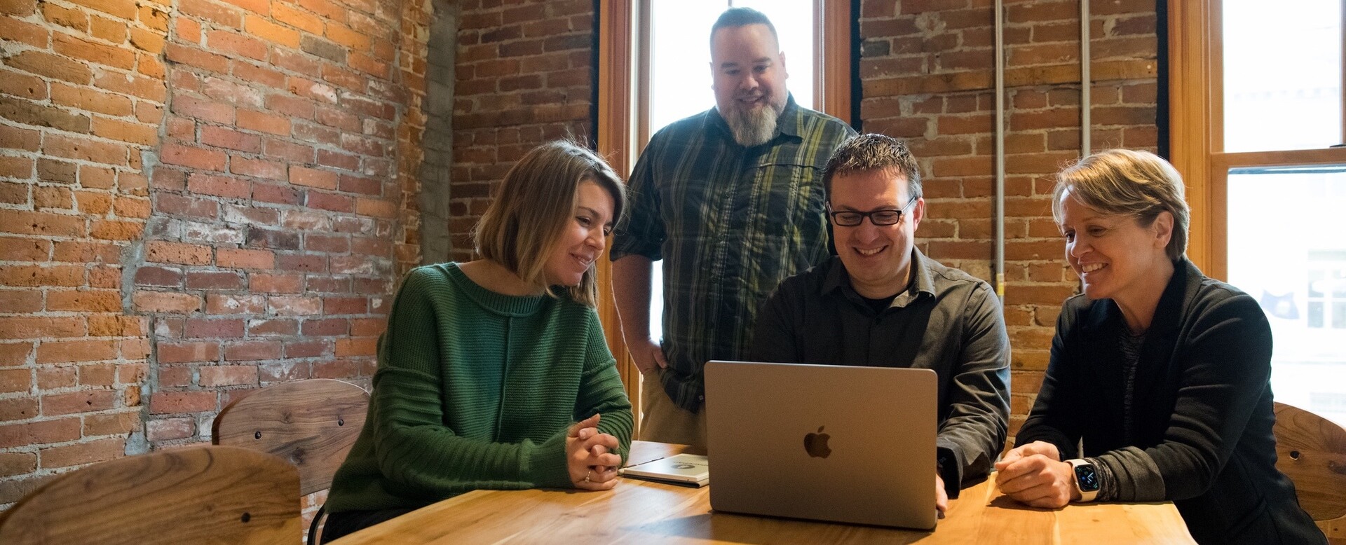 Four people gathered around a laptop, discussing in a cozy, brick-walled setting.