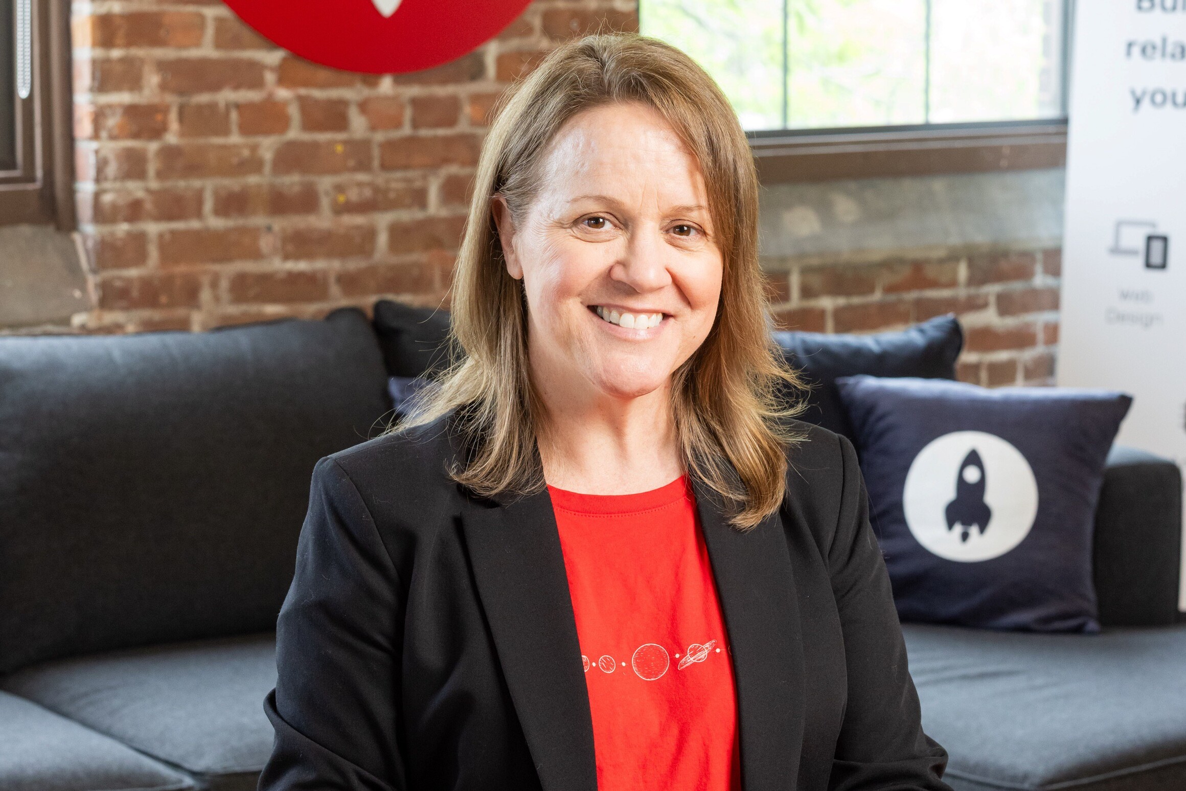 Woman smiling in a black blazer, red shirt, seated on a couch with brick background.
