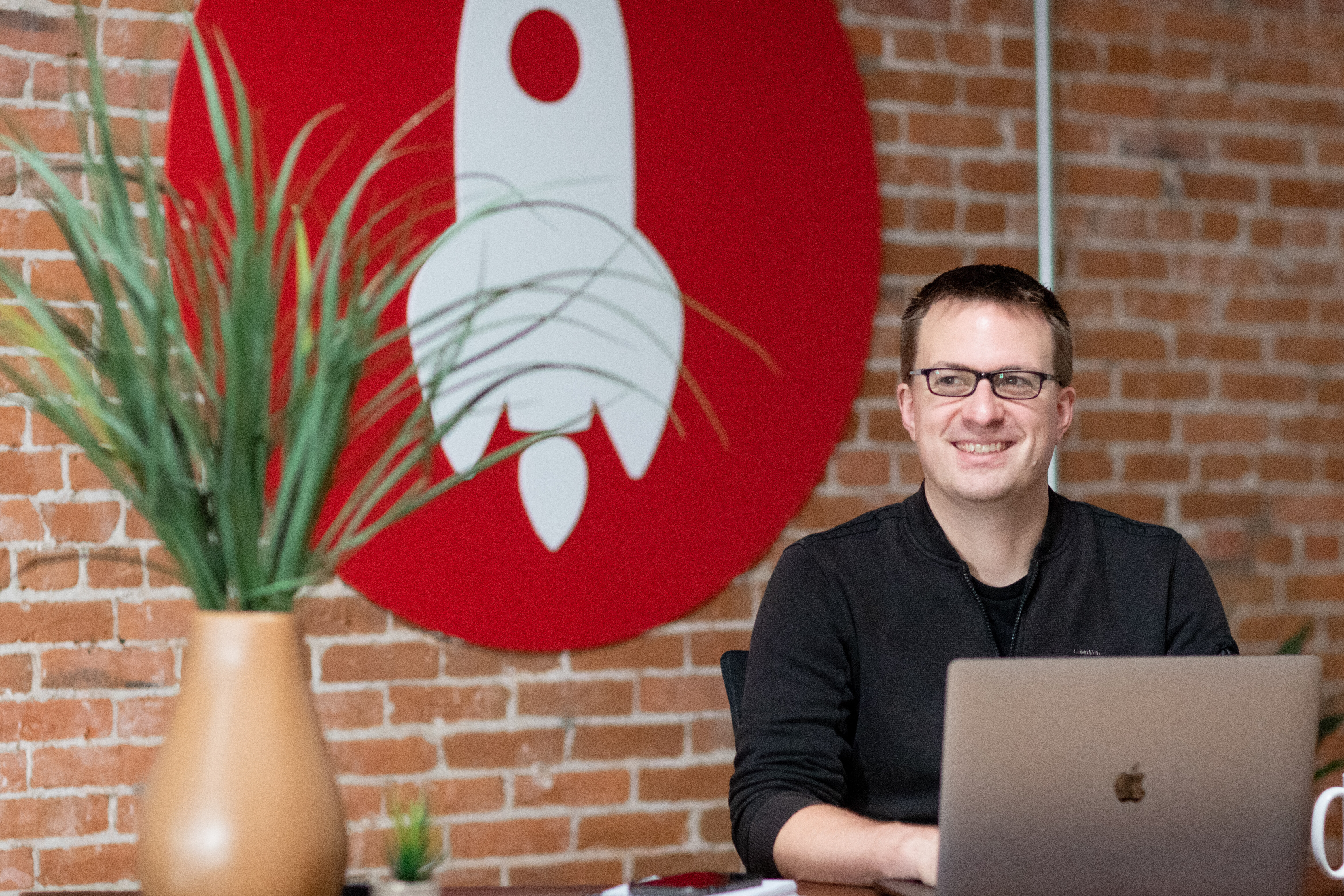 Person smiling at a laptop with a rocket logo backdrop and indoor plants.