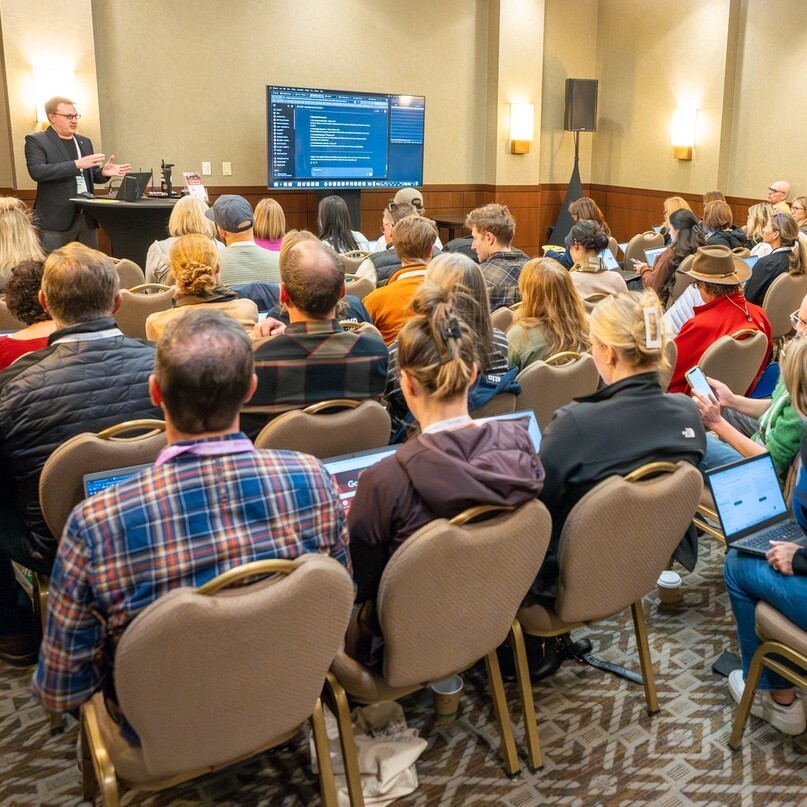 A speaker presents to an audience in a conference room, with people using laptops.
