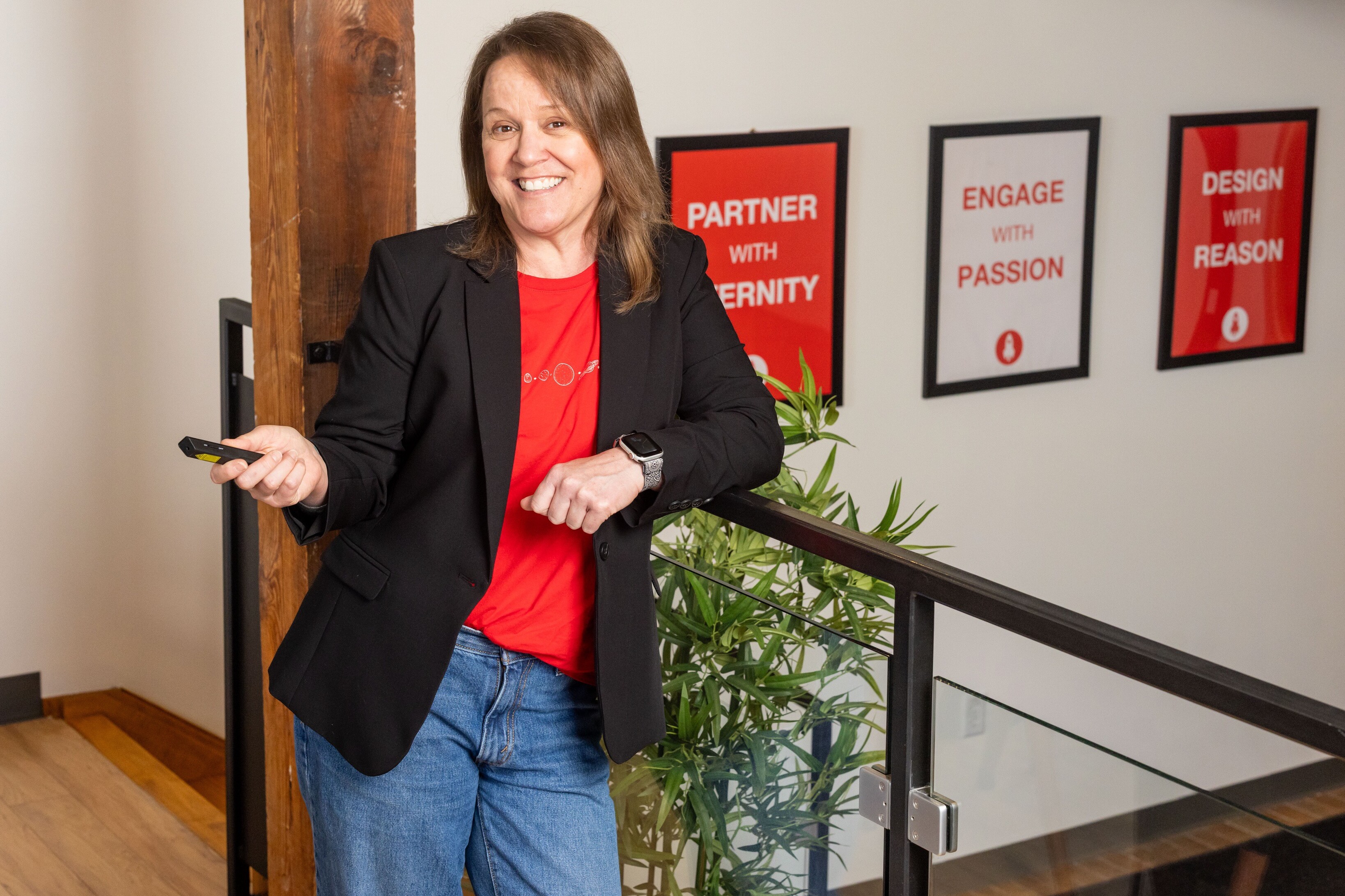 Smiling woman in a blazer and red shirt, standing by a staircase with motivational posters.