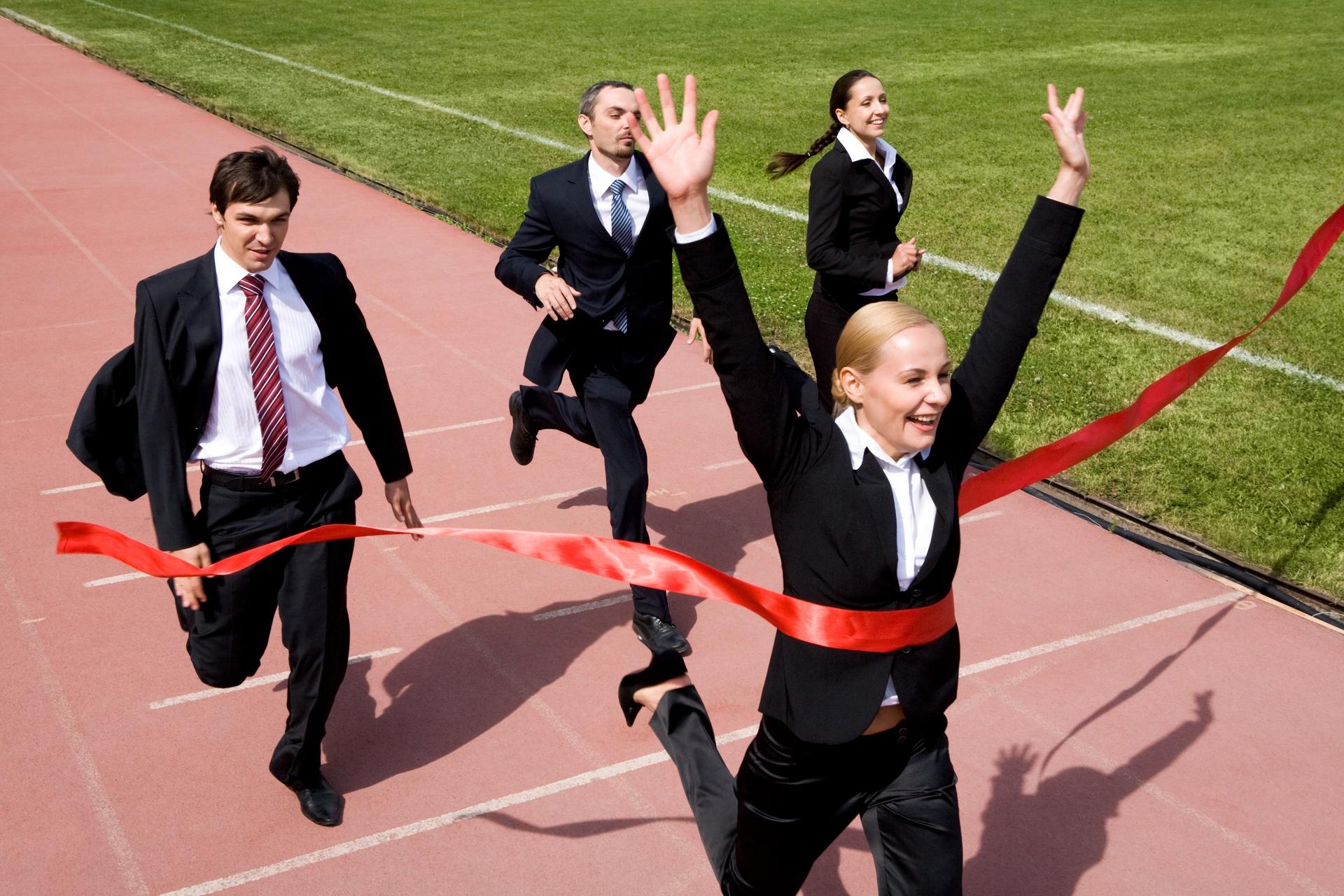 Businesspeople in suits running a race, with one woman breaking the finish line ribbon